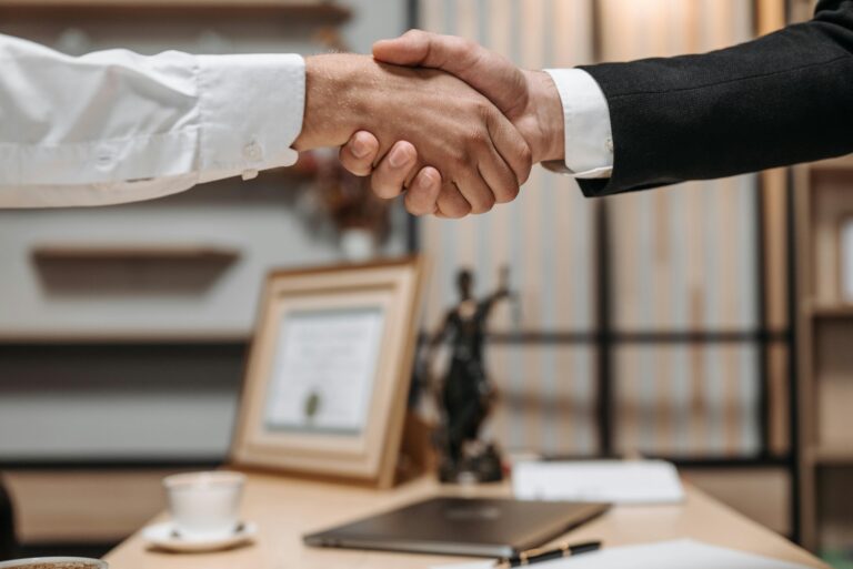 Two professionals shaking hands in an office with the scales of justice in the background, symbolizing legal agreement or mediation settlement.